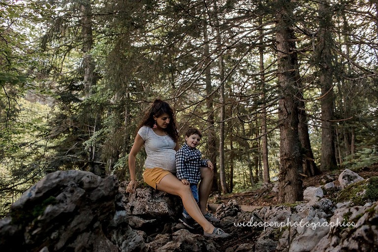 Pregnant woman and young boy sit on rocks in a forest, smiling at the camera.