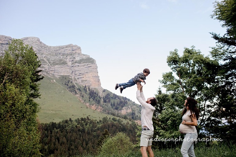 Man lifts a child into the air as a pregnant woman looks on in a mountain meadow.