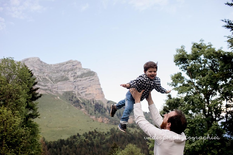 Man lifts a smiling toddler airborne outdoors with mountains and trees in the background, capturing a playful moment.