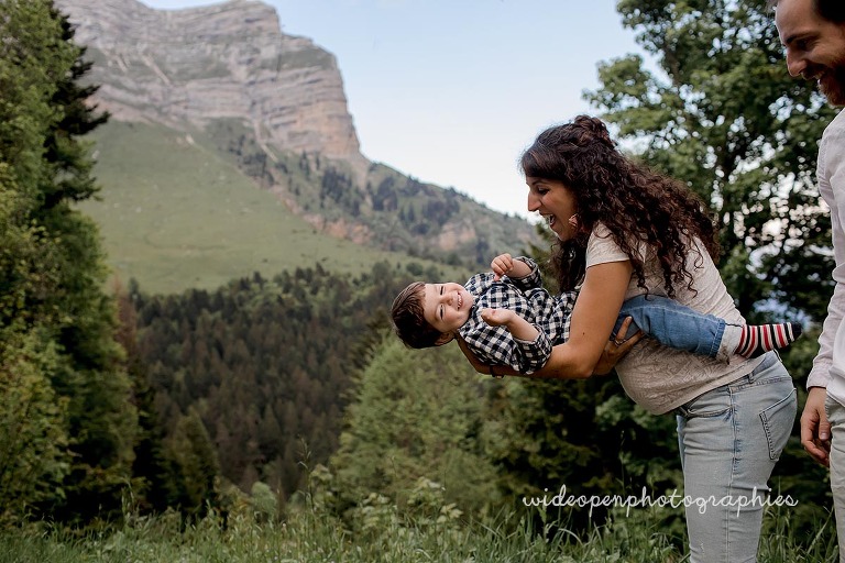 A woman joyfully lifting a smiling child horizontally while a man stands nearby, mountains and trees in the background.