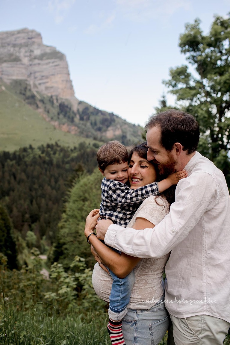 Family of three embracing outdoors in a mountain landscape; expectant mother cradles her baby bump as a young child hugs them.