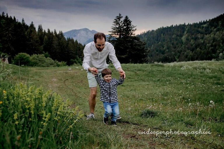 Father helps his young son walk across a grassy meadow with mountains in the background, during an outdoor outing.