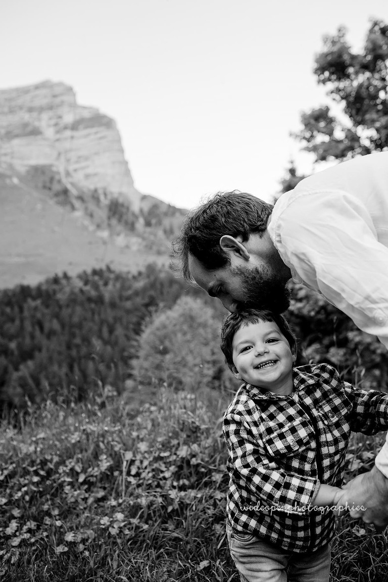 Father leaning down to kiss his smiling toddler in a grassy mountain area outdoors, black-and-white photo.